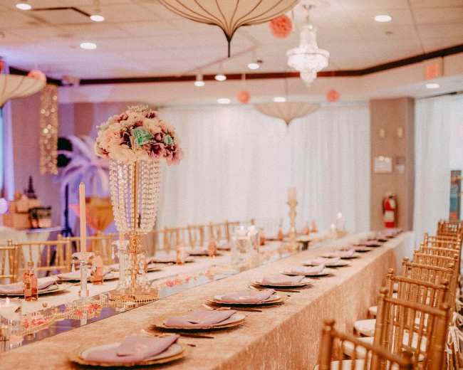 A long banquet table is set with elegant place settings, adorned with a decorative centerpiece and draped in a sparkling cloth under stylish lighting fixtures.