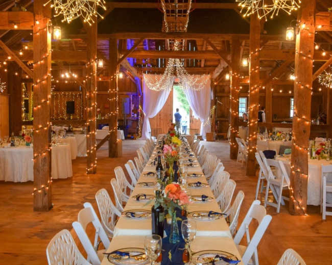 A decorated barn venue with a long dining table set for a celebration, featuring white chairs and string lights overhead.