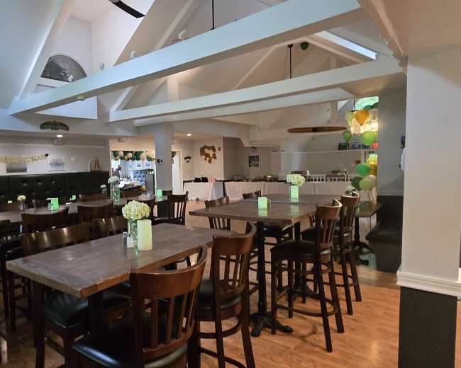 The image shows a spacious restaurant interior with wooden tables and chairs, decorations along the walls, and a designated area for a buffet or event setup.