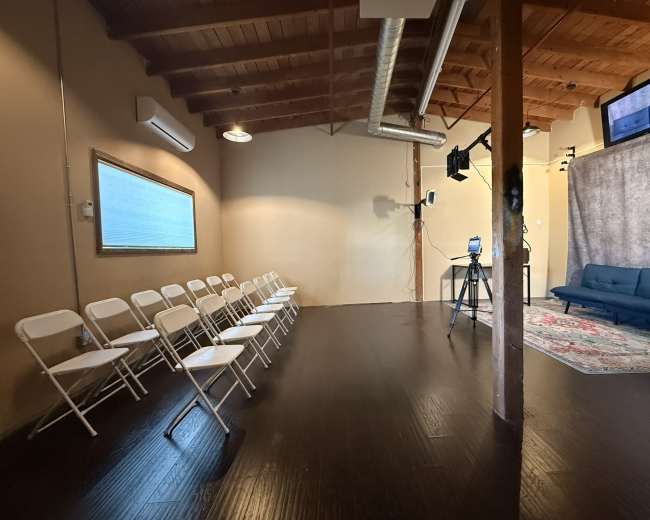 The image shows a sparsely furnished room featuring a row of white folding chairs, a blue couch, and a backdrop with a camera set up for a possible recording or presentation.
