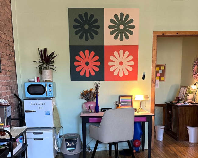 A cozy workspace features a desk with a chair, colorful wall art, a small refrigerator, and a patterned rug on wooden flooring.
