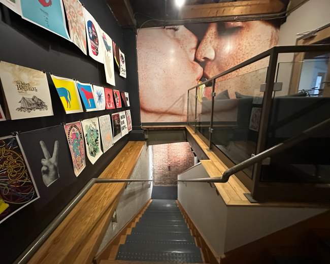 A stairway leading down with wooden steps, flanked by artwork on the walls, and a large mural of a kiss at the end of the corridor.
