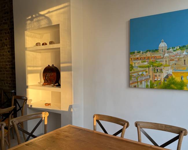 A wooden dining table with cross-back chairs is illuminated by sunlight, alongside a wall featuring a painting and a white shelf displaying decorative items.