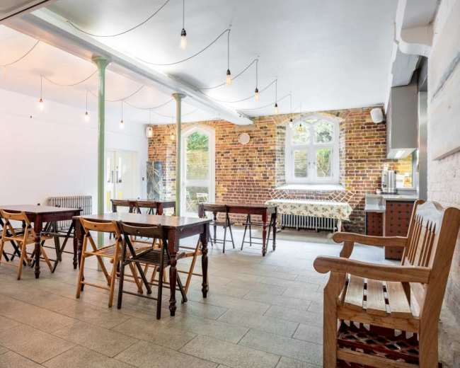A spacious room with wooden tables and chairs, exposed brick walls, and large windows letting in natural light.