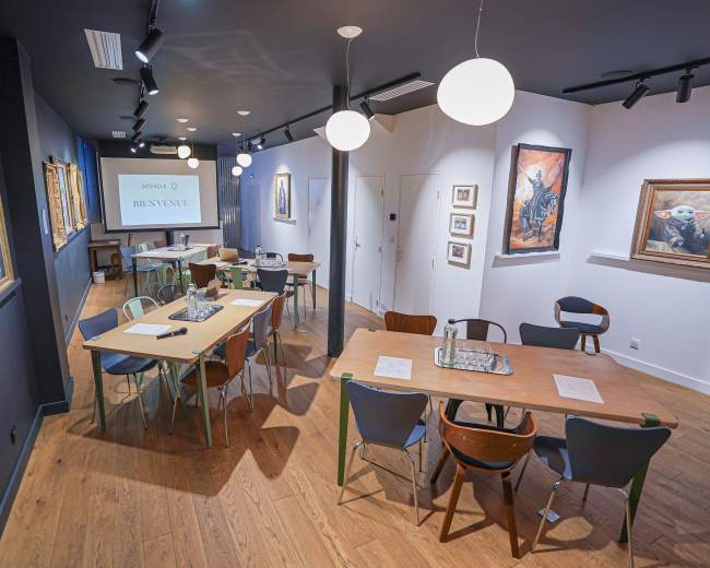 The image depicts a modern, well-lit art studio or gallery space with tables and chairs, framed artwork on the walls, and a screen displaying the word "BIENVENUE."