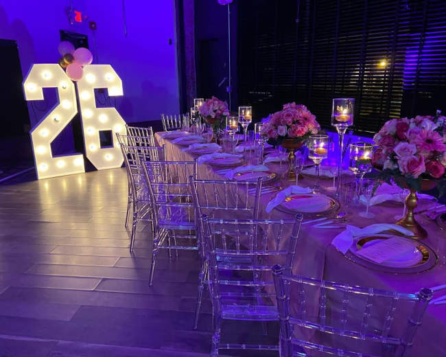 A long banquet table is set with elegant tableware and floral centerpieces beside a large illuminated sign displaying "26."