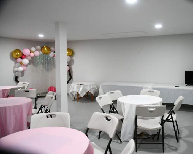 A spacious event room features round tables with pink tablecloths, white chairs, and a decorative backdrop of balloons against a plain wall.