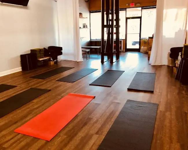 The image shows a yoga studio with several exercise mats arranged on a wooden floor, illuminated by natural light from large windows.
