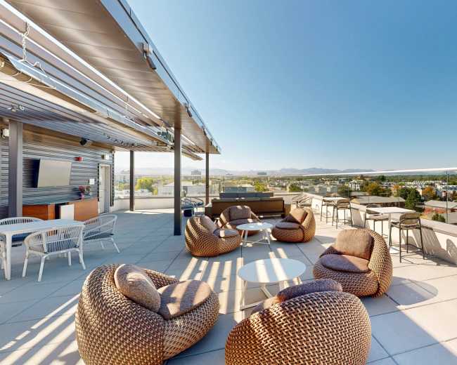 A rooftop terrace features various seating options, including round tables and woven lounge chairs, overlooking a panoramic view of the surrounding landscape.