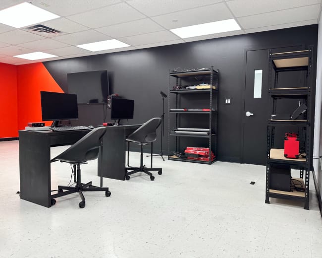 The image shows a workspace featuring two desks with computers, a shelving unit, and a solid black and orange wall.