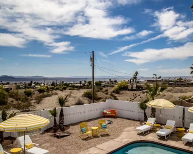 A desert landscape with a swimming pool surrounded by lounge chairs and palm trees, under a blue sky with scattered clouds.