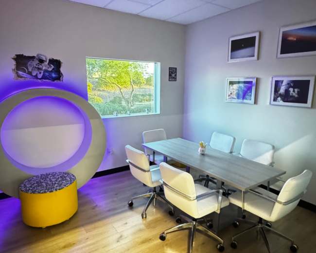 The image shows a modern conference room with a round, illuminated seating area, a wooden table surrounded by white chairs, and large windows with natural light.