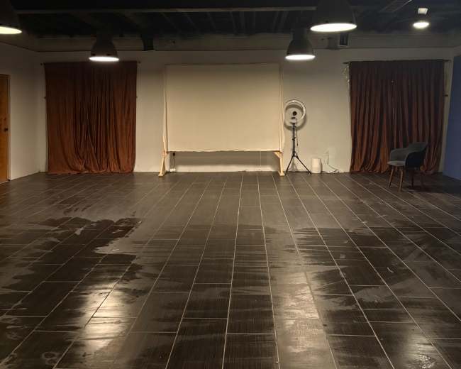 The image shows a spacious room with dark tiled flooring, featuring two curtains on the left, a blank screen in the center, and a single chair in the corner.
