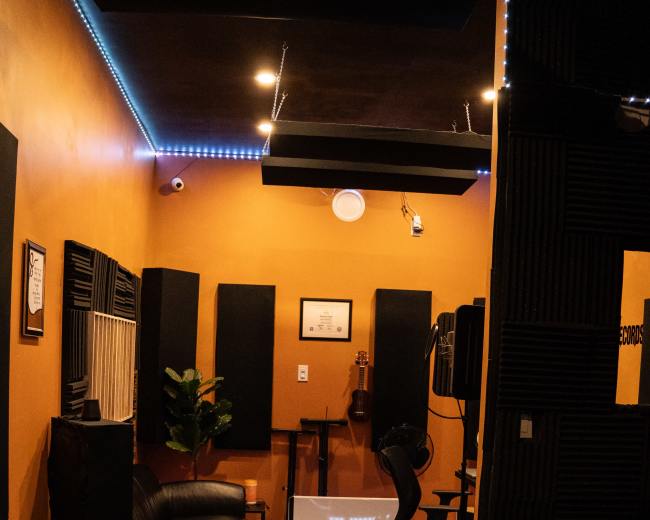 The image shows a recording studio with dark orange walls, black soundproofing panels, seating, and equipment arranged for music production.