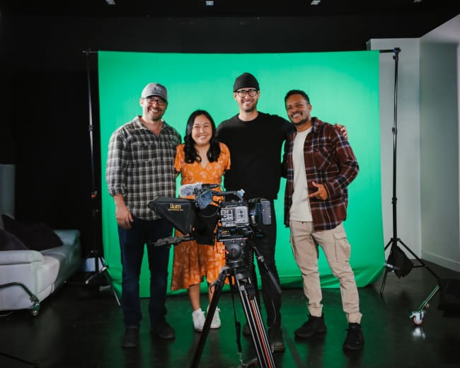 Four individuals pose in front of a green screen, with a camera and lighting equipment set up for a video shoot.