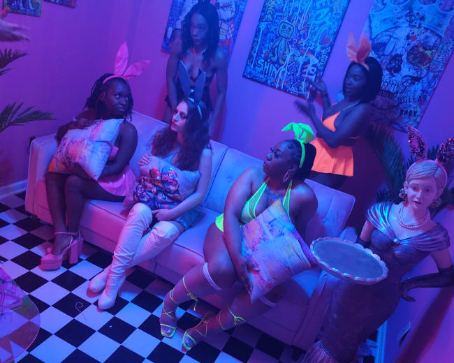 A group of five women, dressed in colorful attire, are seated and standing in a vibrant, neon-lit room with a patterned floor and artwork on the walls.