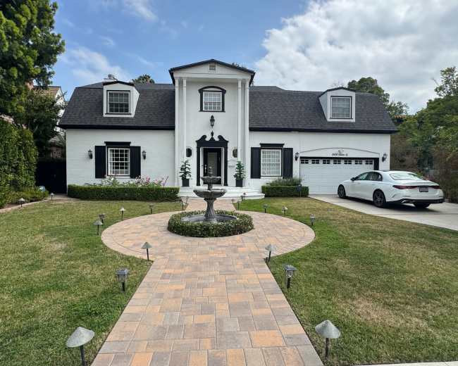 The image shows a two-story white house with a black roof, surrounded by a landscaped yard featuring a central pathway leading to a fountain.