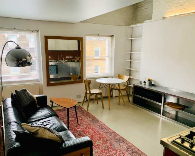 The image shows a modern living area featuring a black leather sofa, a round dining table with chairs, and shelves along the wall in a well-lit room.