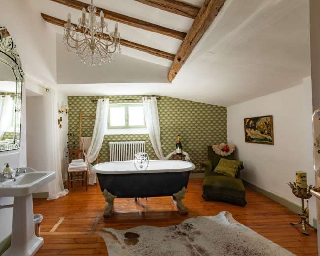 A spacious bathroom features a freestanding bathtub, a chandelier, and vintage decor with wooden beams and a patterned wall.