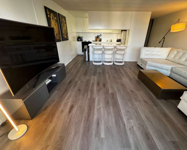 The image shows a modern living room with a large television, a white sectional sofa, a coffee table, and a kitchen area with bar stools in the background.