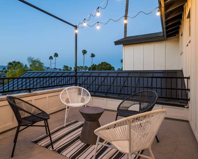 A rooftop terrace features four chairs arranged around a small table, with string lights overhead and palm trees in the background.
