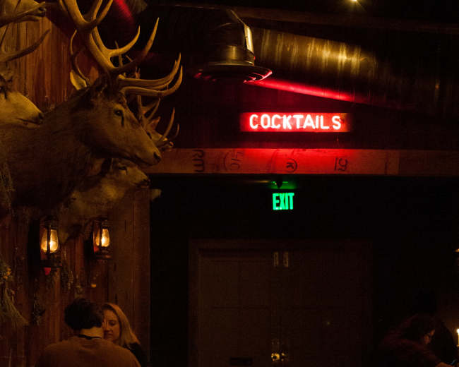 A dimly lit bar interior features a mounted deer head and a glowing red "COCKTAILS" sign above a wooden seating area.