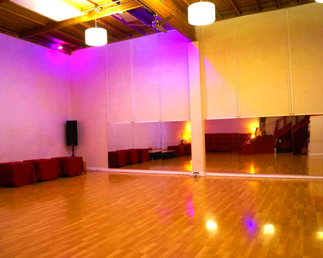 A spacious dance studio with wooden floors, purple lighting, audio equipment, and large mirrors on one wall.