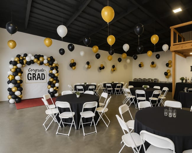 The image shows a decorated event space for a graduation celebration, featuring black and gold balloons, a "Congrats Grad" sign, and tables arranged for guests.