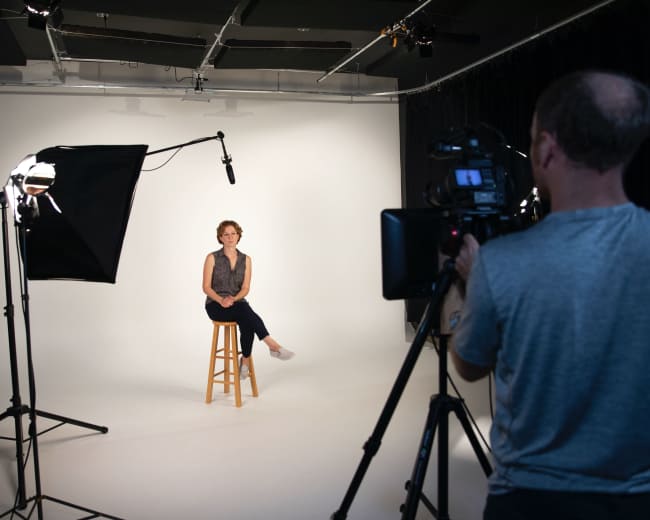 A woman sits on a wooden stool in a well-lit studio while being filmed by a camera operator.