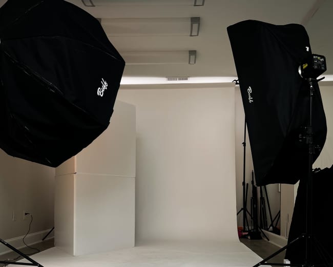 A photography studio setup features two large softbox lights directed at a blank backdrop with a white pedestal.
