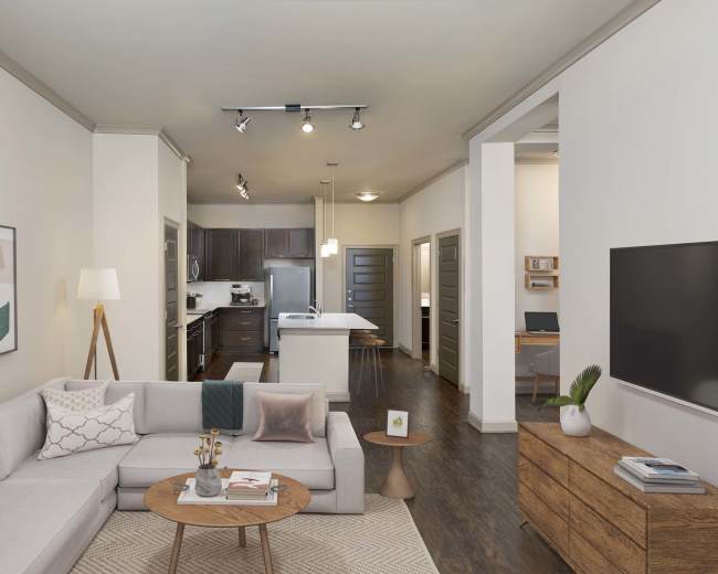 The image shows a modern living room featuring a light gray sectional sofa, a round coffee table, and a television mounted on the wall, with a kitchen visible in the background.