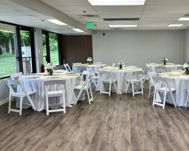 A spacious room features round tables covered with white linens and decorated with small flower arrangements.