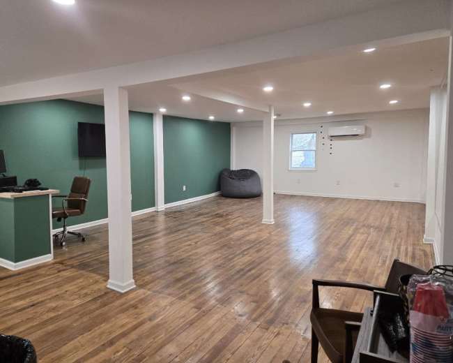 The image shows a spacious room with wooden flooring, multiple lighting fixtures, and a green accent wall, along with a desk and chair, a beanbag, and an air conditioning unit on one side.