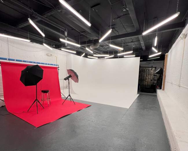 The image shows a photography studio featuring a red backdrop on one side, a white backdrop on the other, and various lighting equipment overhead.