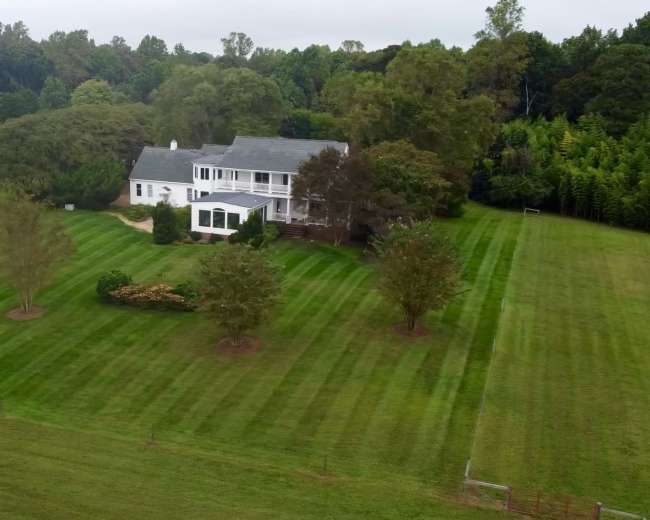 A large, two-story white house is surrounded by neatly striped green lawn and trees on a spacious property.