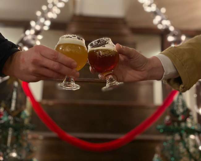 Two hands clink glasses of beer on a staircase adorned with twinkling lights and Christmas decorations.