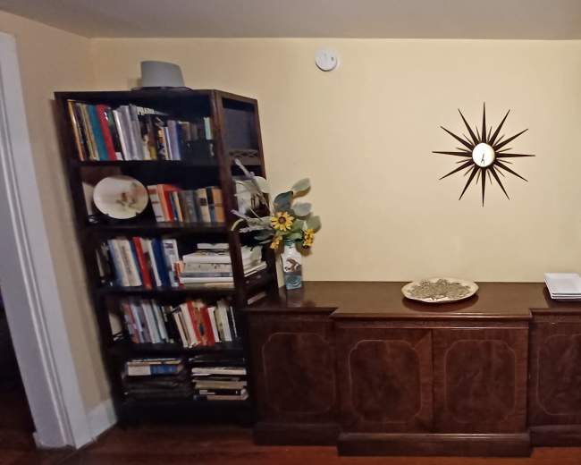 The image shows a furnished corner of a room featuring two bookshelves filled with books, a decorative wall clock, and a wooden cabinet with a tray on top.