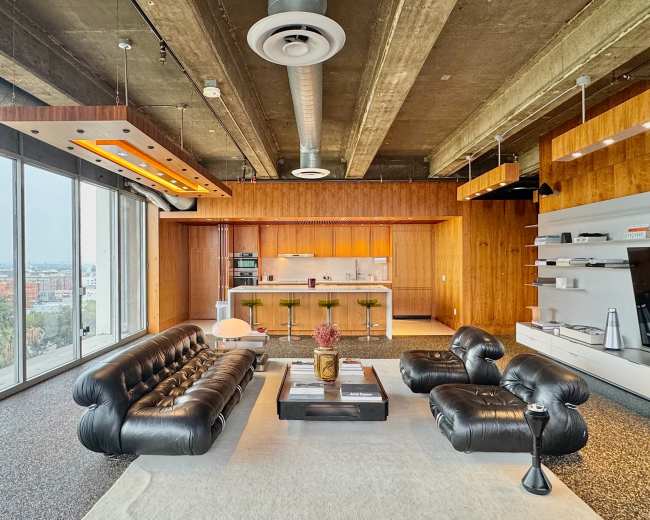 The image shows a modern living space with a black leather sofa, a coffee table, and a kitchen area featuring wooden cabinetry and bar stools.