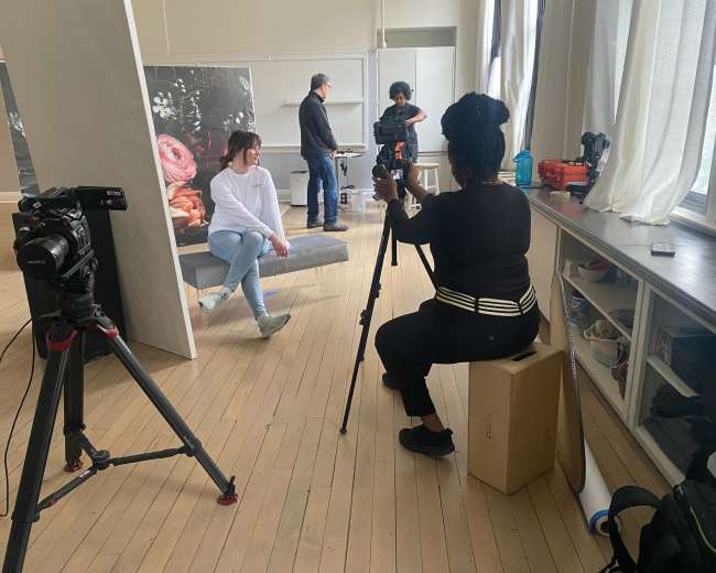 A woman sits in front of a camera while another person poses on a platform in a studio setting.