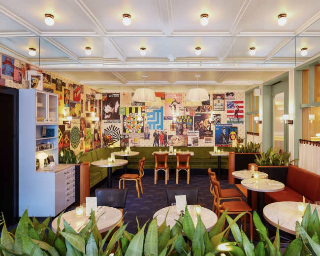 The image shows an interior of a restaurant featuring green and brown seating, round tables, and a collage of colorful posters on the walls.