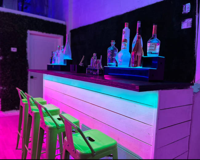 A modern bar with a glowing neon-lit countertop and various bottles of liquor displayed on shelves behind it, accompanied by green metal stools in front.