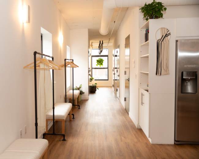 A long, narrow hallway features two clothing racks, benches on either side, and large windows that let in natural light.