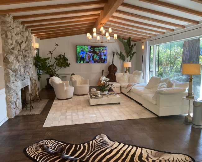 The image shows a modern living room with a stone fireplace, large windows, white furniture, and a patterned area rug.