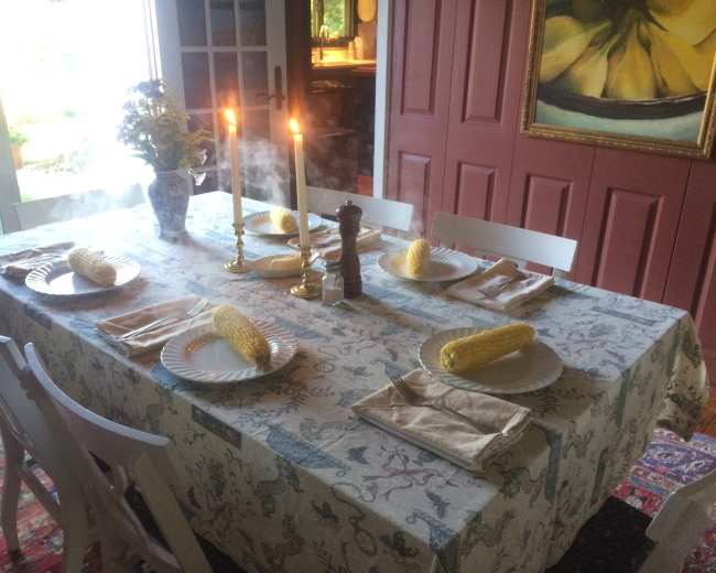 A dining table is set with plates of corn, utensils, and lit candles, surrounded by chairs and decorated with a floral vase.