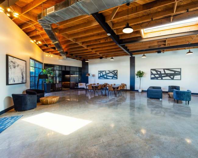 The image depicts a spacious, modern interior with high wooden ceilings, polished concrete flooring, several seating areas, and large windows allowing natural light to enter.