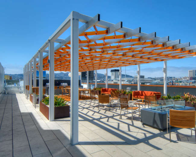 The image shows a rooftop terrace featuring a pergola with seating areas and planters, overlooking a city skyline.