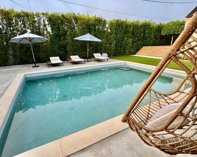 The image shows a modern swimming pool surrounded by lounge chairs and umbrellas, with a neatly trimmed hedge and grass area in the background.