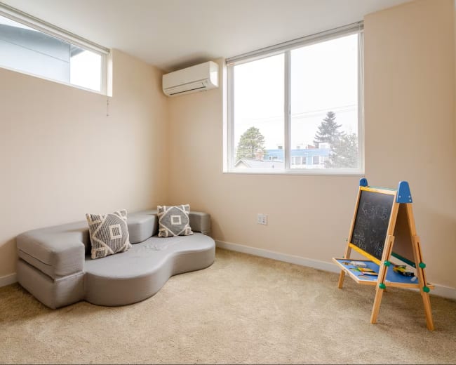 The room features a gray sectional sofa, two decorative pillows, and a blackboard easel near a large window.