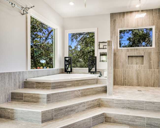 The image shows a spacious bathroom featuring a large tub on a raised platform, a walk-in shower, and large windows that provide a view of outdoor greenery.