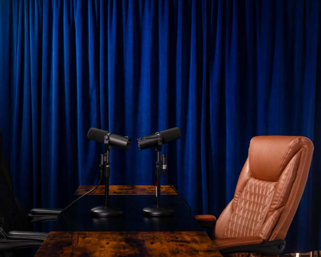 Two office chairs, one black and one brown, are positioned opposite each other at a table with two microphones, set against a blue curtain backdrop.
